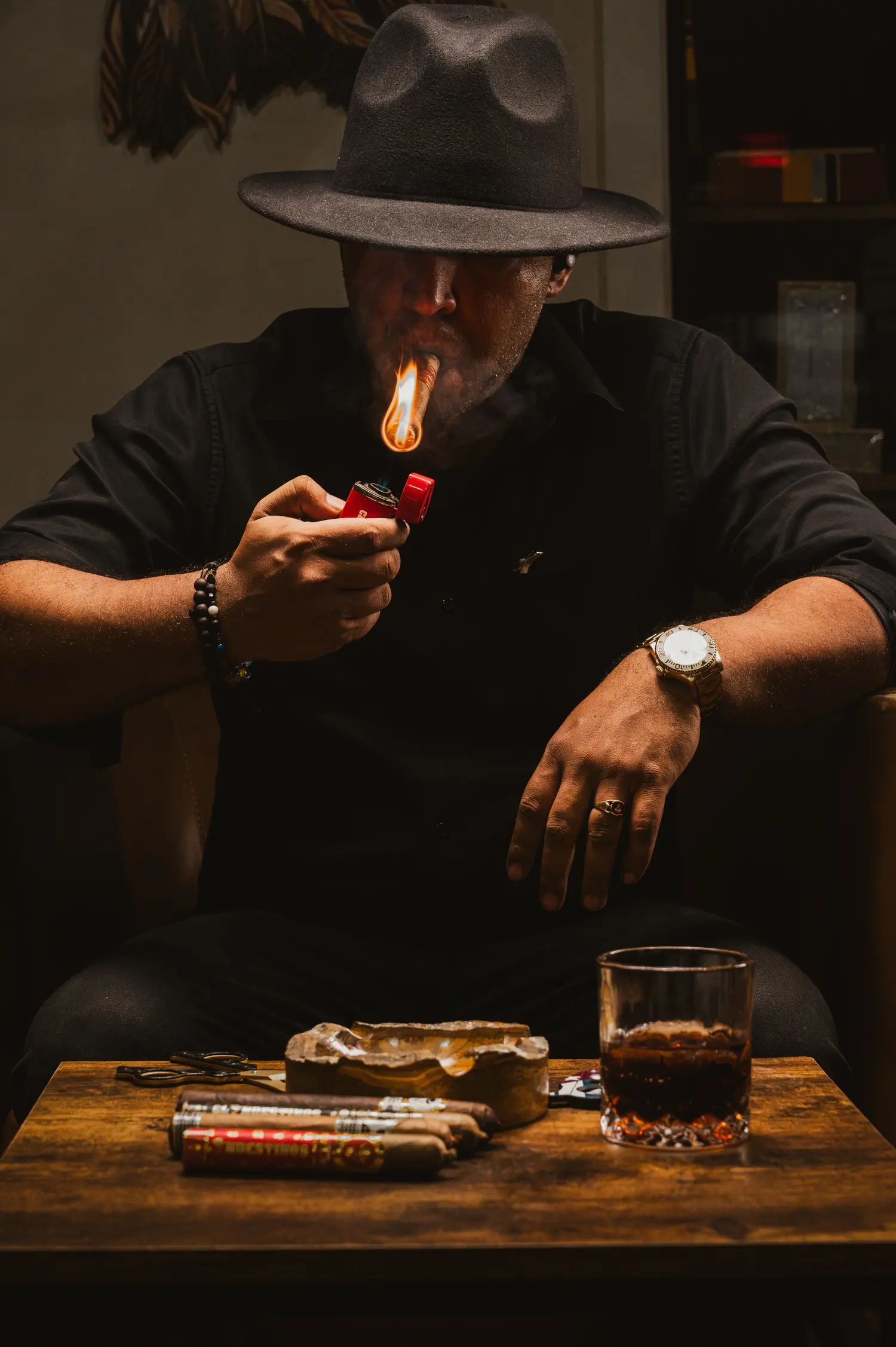 A man lighting a cigar at a table with a glass of whiskey