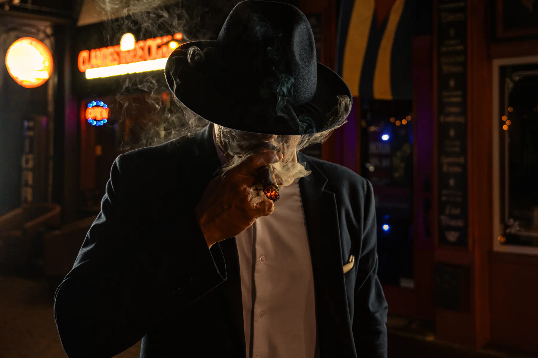 A man in a suit and hat outside the Clandestinos boutique at night, his face hidden by cigar smoke